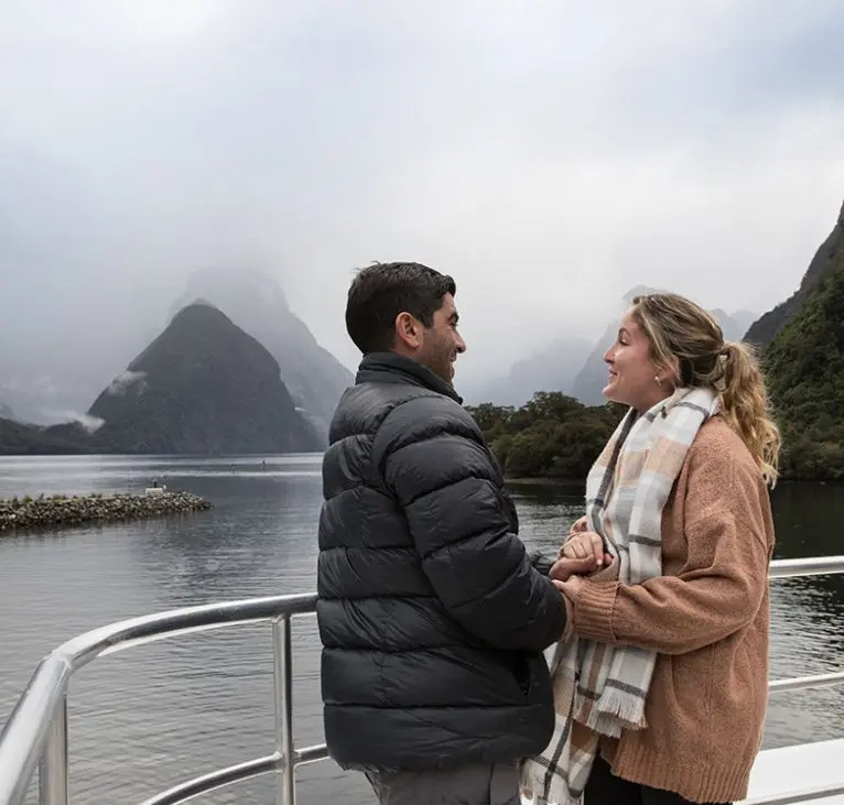 A couple standing on the deck of a tour boat in Milford Sound, dressed in winter clothes.